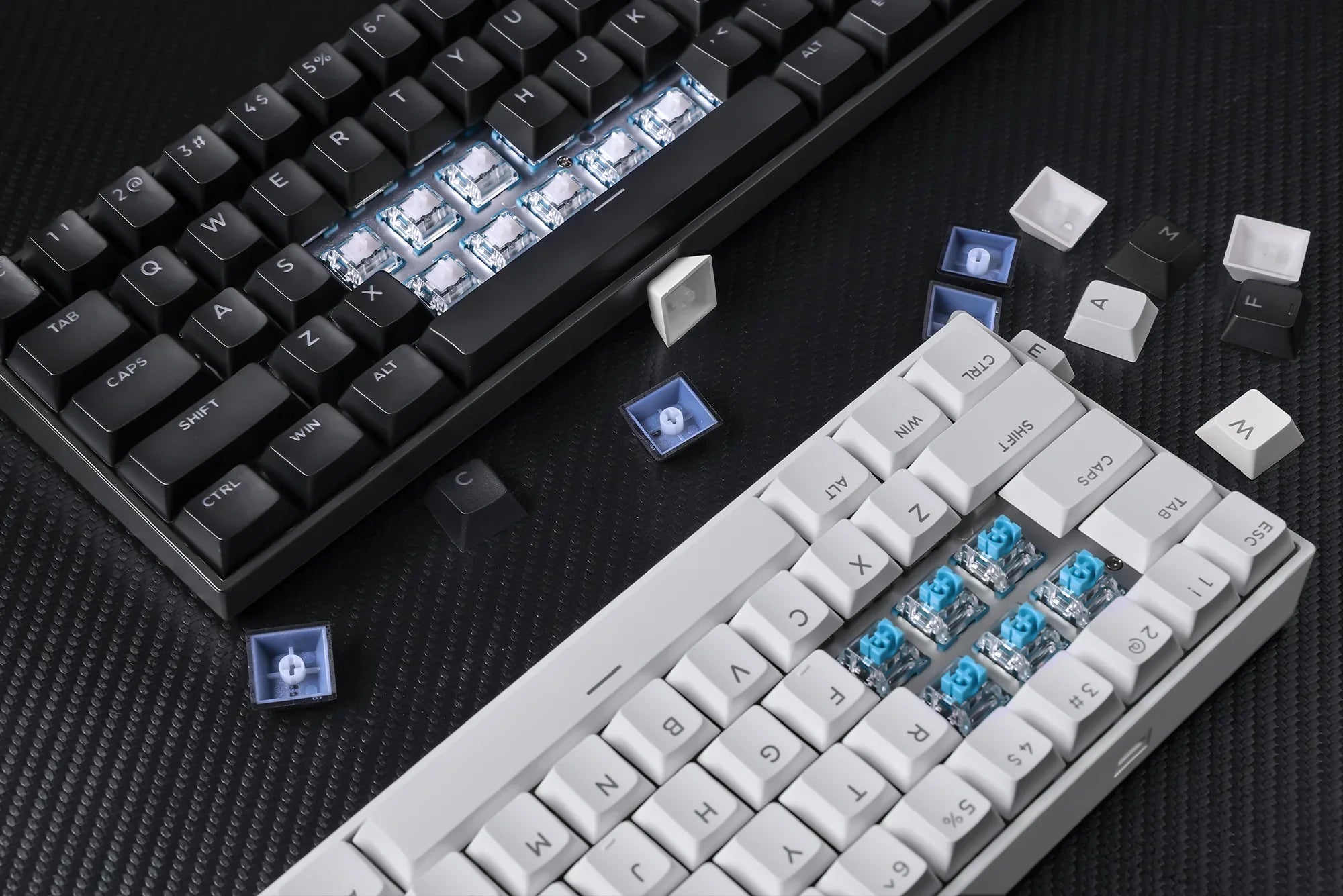 Black and white mechanical keyboards with exposed blue switches and scattered keycaps on textured surface